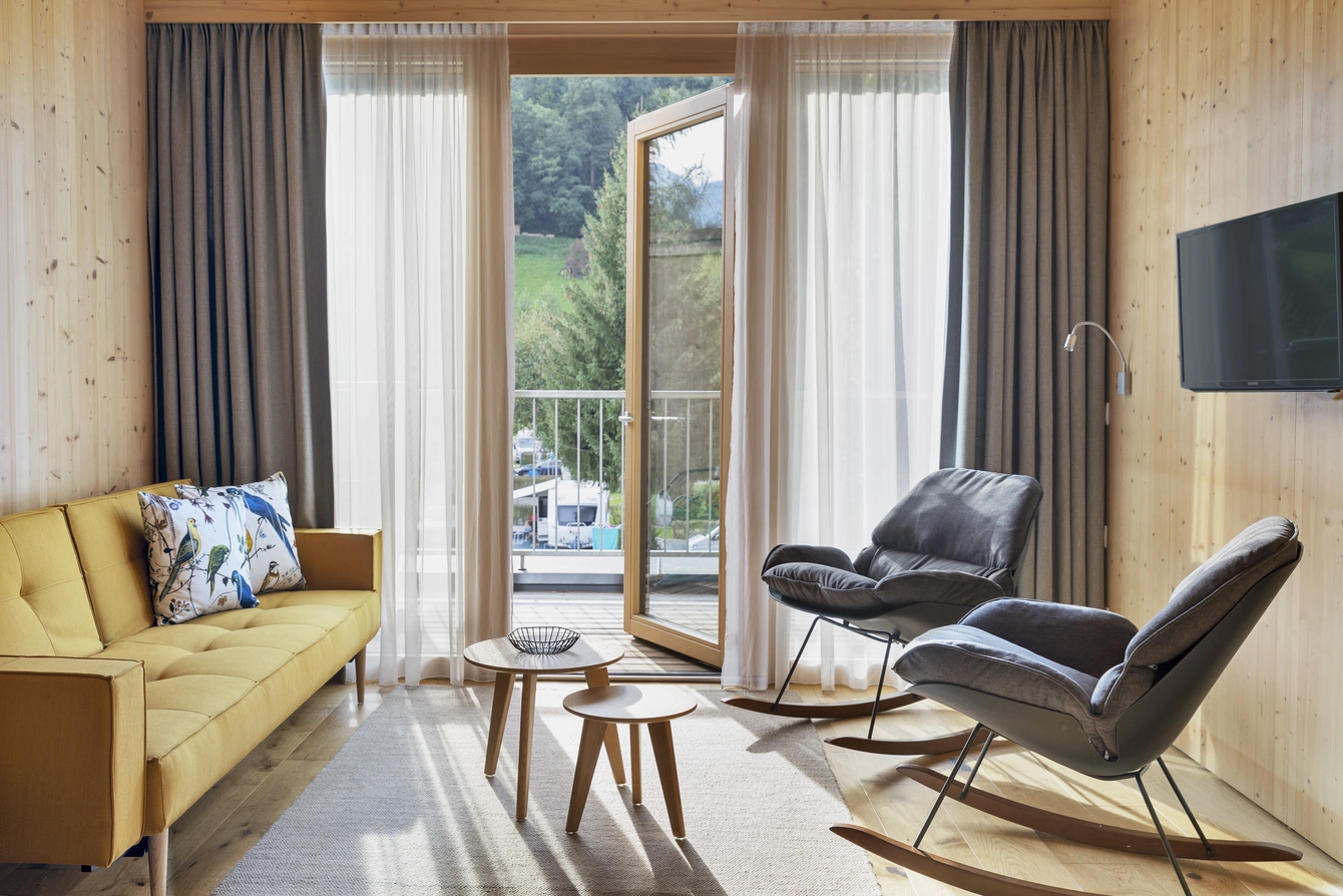 Apartment Sonnwend ©Natürlich HELL Camping & Aparthotel Der Wohnbereich des Sonnwend Apartments mit einem gelben Sofa, einem TV und gemütlichen Stühlen welche einen kleinen Tisch umringen.