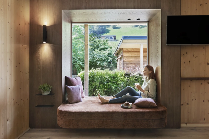 Helles & modernes Apartments ©Natürlich HELL Camping & Aparthotel Eine Frau die in der gemütlichen Sitzecke sitzt, welche mit Polstern dekoriert ist und Blick auf den Garten bietet.