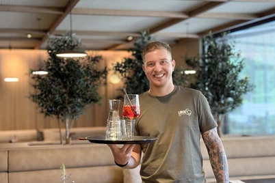Ein Kellner steht lächelnd im Restaurant und hält ein Tablett mit Getränken in einer Hand. Im Hintergrund sind die Sitzbänke, Tische und Pflanzen zu sehen.