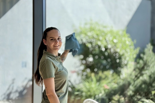 Eine Mitarbeiterin des Sauber-Teams während sie die Fensterfront putzt und mit einem Lächeln über die Schulter blickt.