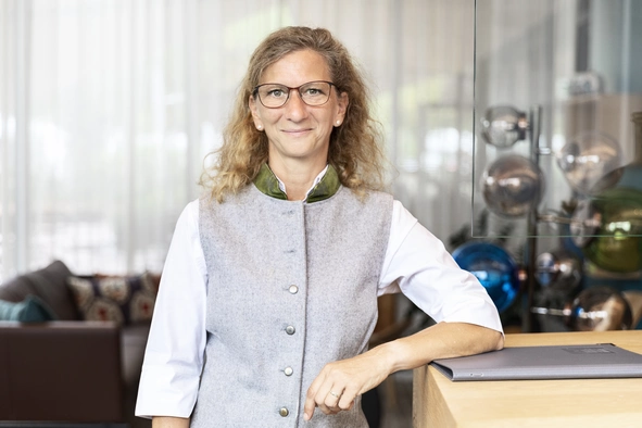 Eine Rezeptionsmitarbeiterin mit blonden locken und der Dienstkleidung die aus einem hellgrauen Gele und einer weißen Bluse besteht mit einem Arm an die Rezeptionstheke anlehnt.