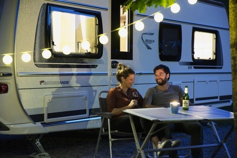 Ein Paar das am Abend vor dem Camper sitzt welcher außen mit einer Lichterkette dekoriert ist.