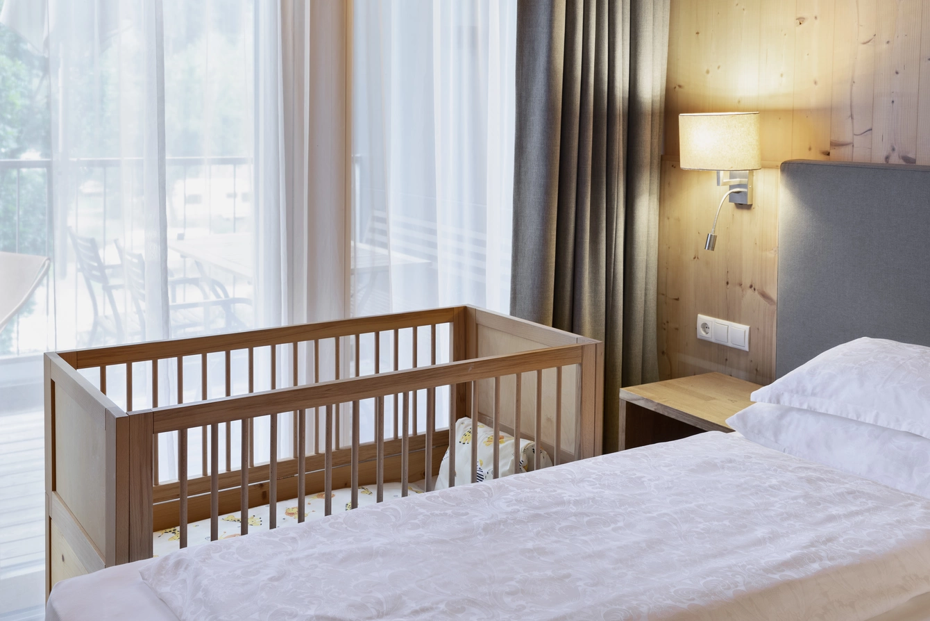 Apartment Sonnwend ©Natürlich HELL Camping & Aparthotel Ein Babybett welches neben dem mit weißer Bettwäsche bezogenem Doppelbett platziert ist.