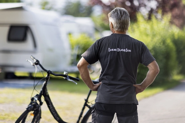 Der Hausmeister mit dem Rücken zur Kamera und einem angelehnten Fahrrad wie er vor dem Campingplatz posiert.